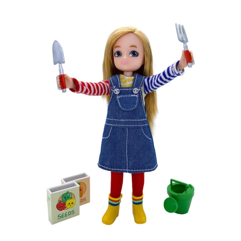 Garden Time Lottie - Doll standing with blonde hair, denim overalls dress, striped shirt. She's holding garden tools and is standing next to seed boxes and a green watering can