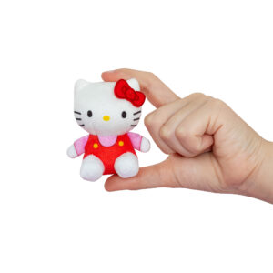 A hand holding a small Hello Kitty plush toy in a red outfit and pink shirt with a red bow on its head against a white background.