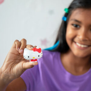 A child with long dark hair, wearing a purple shirt, smiles while holding up a small Hello Kitty figurine. The figurine has a white face, red bow, and two black eyes with whiskers. The blurred background decor includes star shapes.