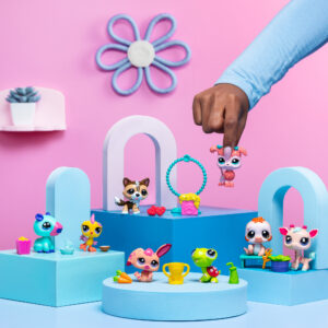 A playful scene featuring several 'Littlest Pet Shop' toy figures arranged on pastel blue and pink geometric platforms against a pink background. A hand is holding one of the pet figures, a pink dog, in mid-air. Other toys include a blue and pink creature with a yellow drink, a yellow bird with a blue cup, a brown dog with red accessories, a green turtle with a trophy, a pink bunny with a carrot, a white and orange puppy, and a white and pink deer-like figure. The background is decorated with a small plant on a shelf and a flower-shaped wall decoration.