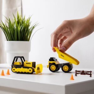 A hand playing with yellow Tonka toy construction vehicles on a white surface. One vehicle, with a scoop, faces forward, while the other has its dump bed raised. Small orange cones and a miniature fence are also on the surface. A green potted plant is in the background.