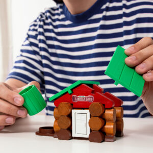 A person wearing a blue and white striped shirt is assembling a small toy log cabin with the brand name "Lincoln Logs" on the red roof. They are holding green pieces that resemble a cylindrical chimney and part of the roof. The cabin has a white door.