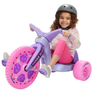 A young girl with curly hair, wearing a helmet, a colorful long-sleeve sweater, and pink pants, is happily riding a purple and pink tricycle with a large front wheel and pink details. She is smiling and appears to be enjoying herself.