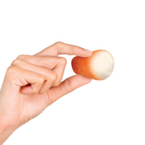 A hand holding a small, round ball that is half tan and half white against a plain white background. The ball looks like a donut hole and is being squeezed. You can see a white filling inside through the material.