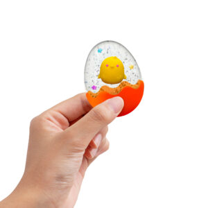 A hand holds a transparent egg-shaped object with a small yellow chick inside. The bottom half of the egg is orange, resembling a cracked shell. Glitter and colorful stars decorate the inside, adding a playful touch. The background is plain white.