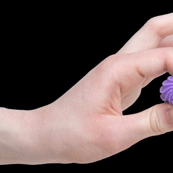 A hand is squeezing a small, purple, textured ball between two fingers. The ball's surface features wavy ridges and is called a Teenie NeeDoh Ripples. The background is plain white.