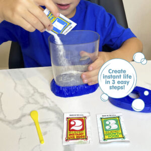 A child wearing a blue shirt is holding the Magiquarium with one hand, and pouring powder from a small packet labled with the number 1 with the other hand. The lid of the aquarium, a packet with the number 2, a packet with the number 3, and a yellow plastic spoon all lay on the surface in front of the child.